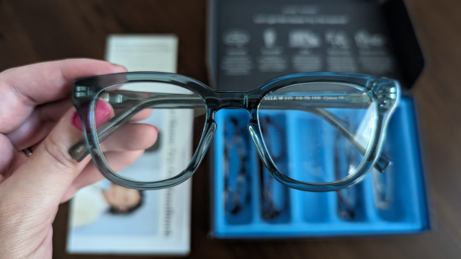 a close-up of a person holding a pair of green warby parker glasses above its home try-on kit